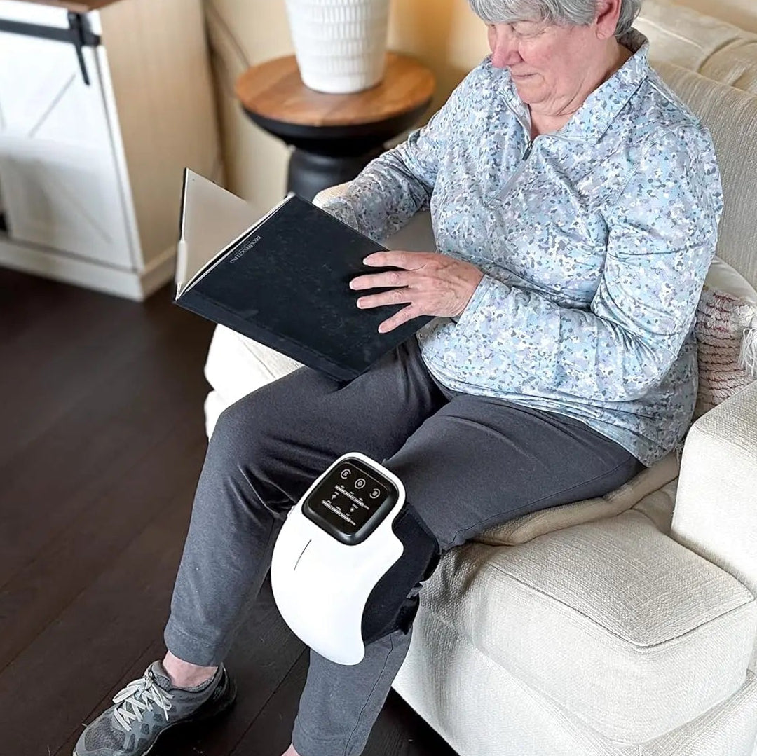 Woman sitting on a couch using a massage device on her leg while reading a book.
